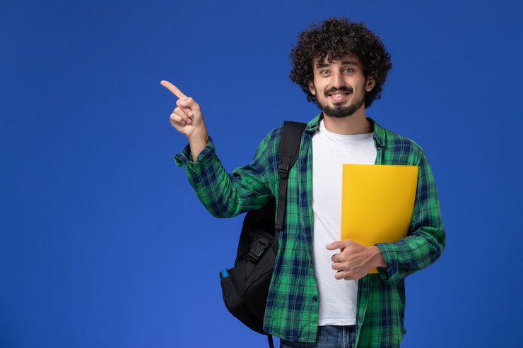 Foto gratuito vista frontale dell'allievo maschio in camicia a scacchi verde che porta zaino nero e che tiene i file sorridenti sulla parete blu